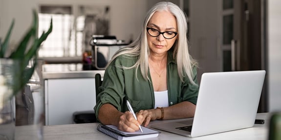 Senior woman taking notes in notebook while using laptop at home. 