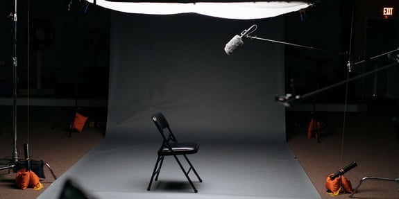 Dark studio with ceiling spotlight shining on an empty chair and microphone in the middle of the room.